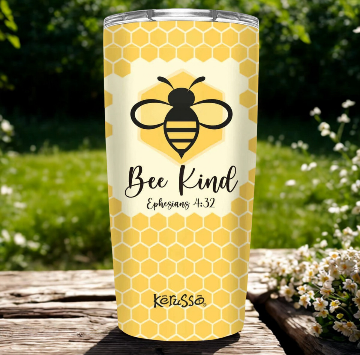 A yellow and white honeycomb-patterned tumbler with a black bee graphic and the text 'Bee Kind Ephesians 3:32' and 'KetUsS' stands on a wooden deck outdoors with green grass and white flowers in the background.