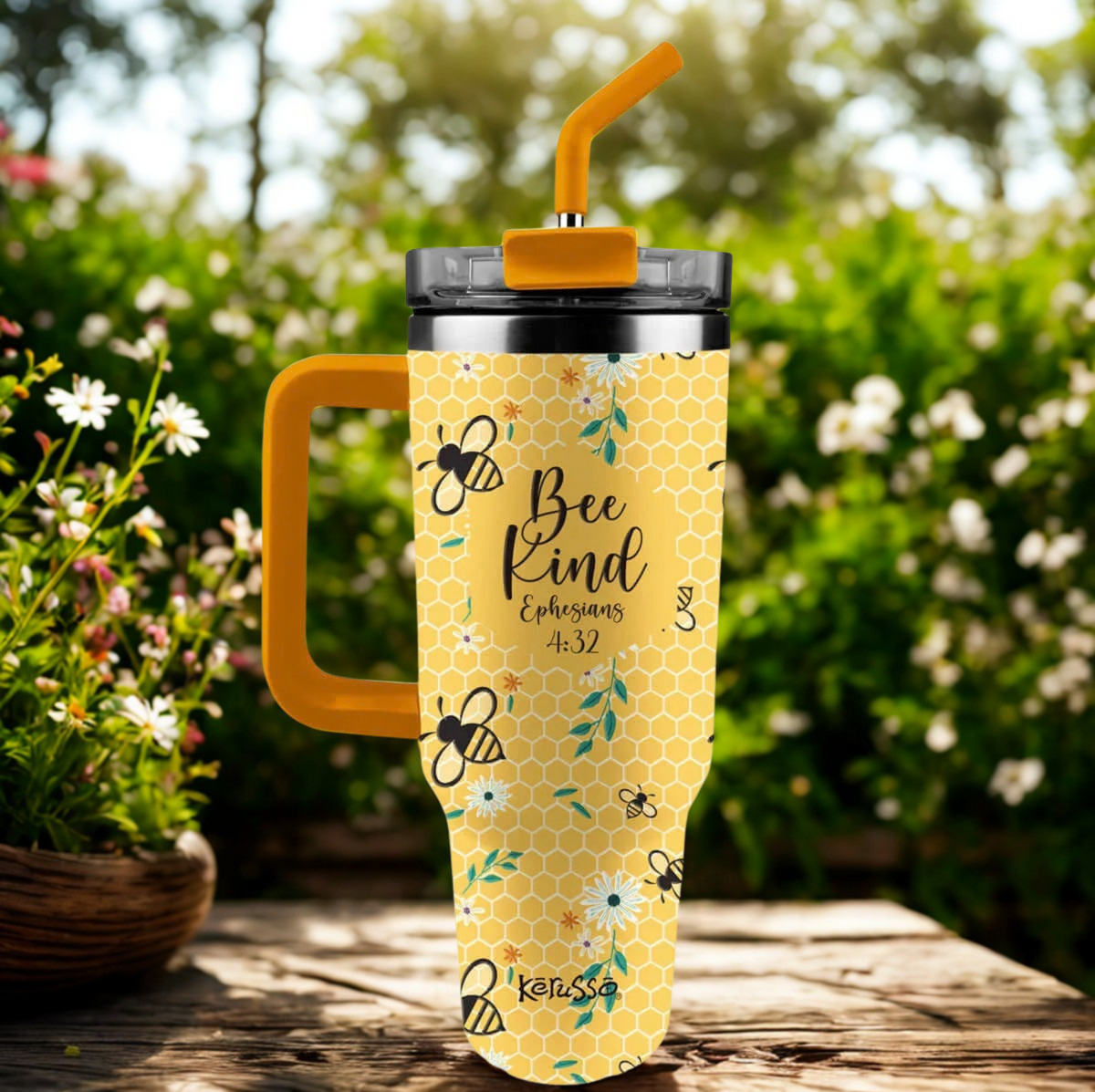 A yellow insulated tumbler with a honeycomb pattern, printed with bumblebees and the text 'Bee Kind Corinthians 16:14', featuring a bright yellow handle and straw, set on a wooden table with a blurred garden background.