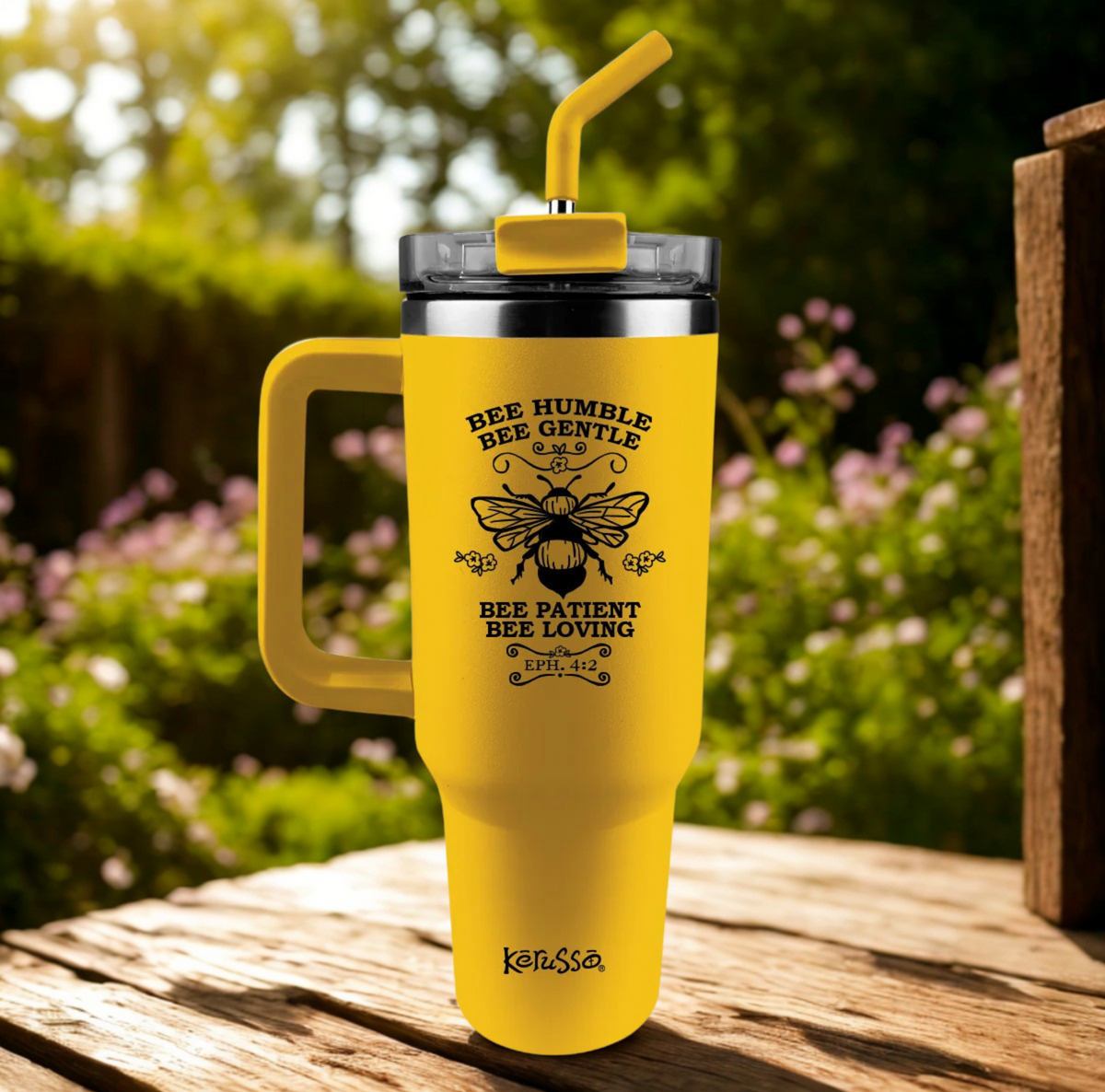 A bright yellow insulated tumbler with a handle, black lid, and yellow straw, standing on a wooden table outdoors. The tumbler has a black bee graphic with the text 'BEE HUMBLE BEE GENTLE BEE PATIENT BEE LOVING' and the brand name 'KARSA' near the bottom.