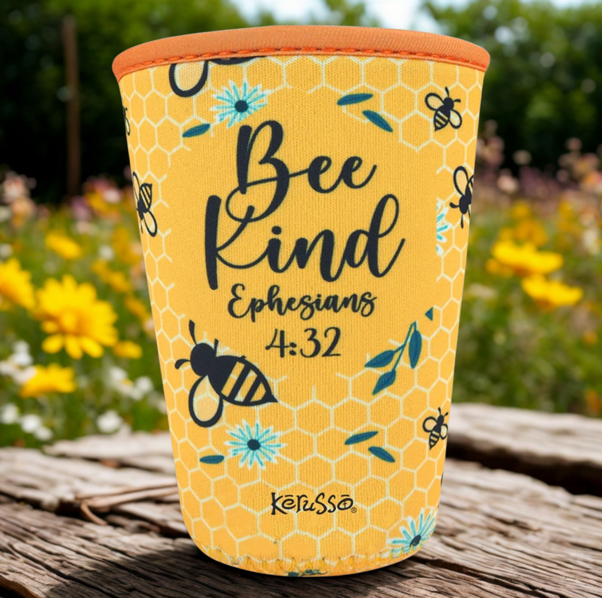 A yellow neoprene cup with a honeycomb pattern, featuring the text 'Bee Kind Ephesians 4:32' and illustrations of bees and flowers. The cup has an orange rim and is shown sitting on a rustic wooden surface with a blurred garden background.