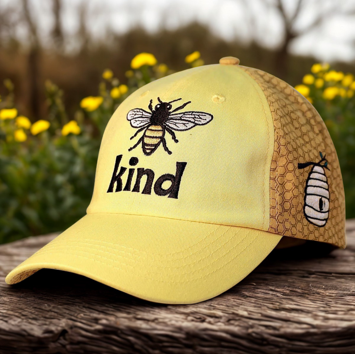 A yellow baseball cap with a honeycomb-patterned back panel, featuring an embroidered bee and the word 'kind' on the front, displayed on a rustic wooden surface with blurred yellow flowers in the background.