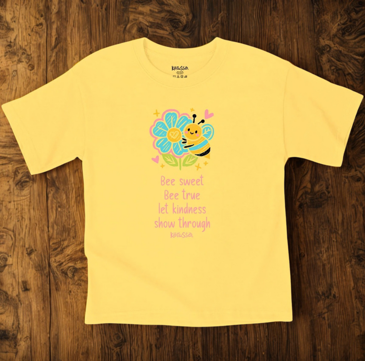 A yellow short-sleeve kids' t-shirt with a colorful bee and flower graphic on the chest, laid flat on a dark wood background.